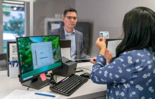 A female bank teller checks the personal identification of a male customer who is seeking to withdraw money from his bank account. Selective focus on the woman's hand as she holds the man's driver's license.